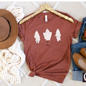 May include: Rust-colored t-shirt featuring three whimsical ghost illustrations with the word "boo" below. The shirt is displayed with a brown hat, cream-colored scarf, blue jeans, and brown flats, suggesting a fall or Halloween theme.