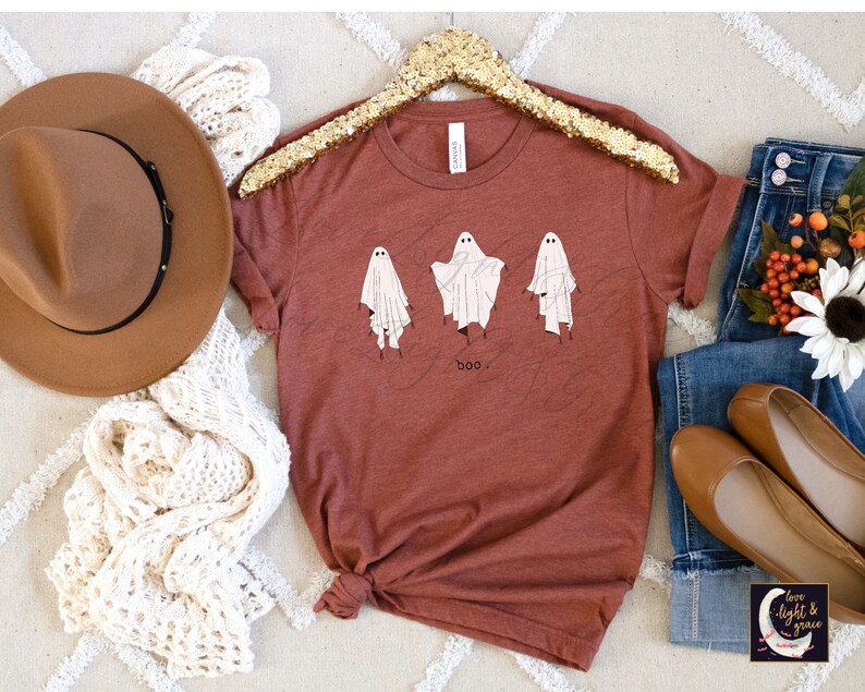 May include: Rust-colored t-shirt featuring three whimsical ghost illustrations with the word "boo" below. The shirt is displayed with a brown hat, cream-colored scarf, blue jeans, and brown flats, suggesting a fall or Halloween theme.