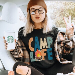May include: Black t-shirt with the words "CAMP LIFE" in colorful, distressed lettering and a campfire graphic. The person is wearing a brown beanie, glasses, and a plaid shirt. A coffee cup with a green logo is held in the left hand.