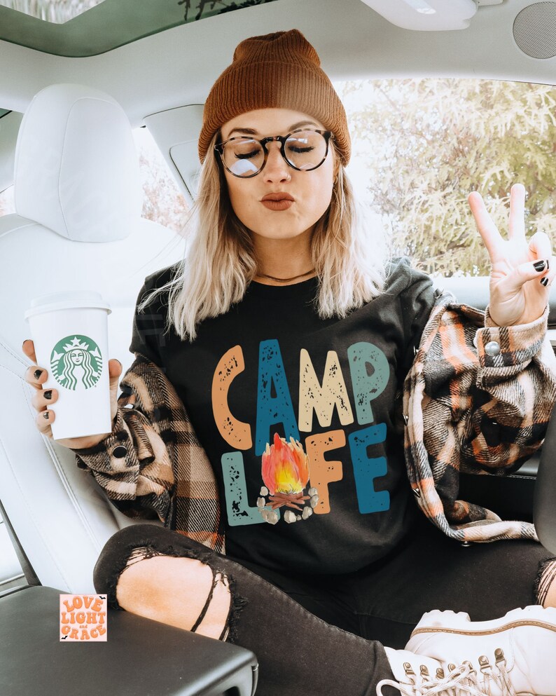 May include: Black t-shirt with the words "CAMP LIFE" in colorful, distressed lettering and a campfire graphic. The person is wearing a brown beanie, glasses, and a plaid shirt. A coffee cup with a green logo is held in the left hand.