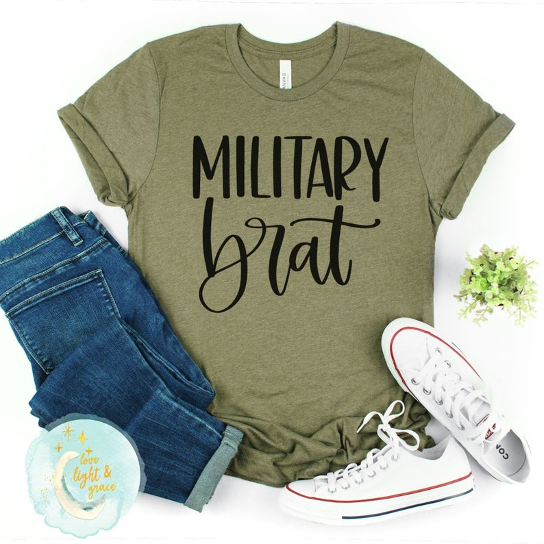 May include: A green t-shirt with the text "MILITARY brat" in black lettering. The shirt is laid flat on a white surface with a pair of blue jeans and white trainers.