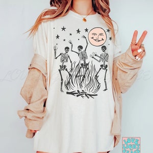 May include: A white t-shirt with a graphic of three skeletons dancing around a bonfire with a smiling moon and stars in the background. The text "Love Light and Grace" is in the bottom right corner.