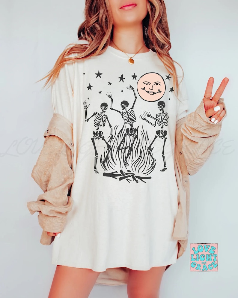 May include: A white t-shirt with a graphic of three skeletons dancing around a bonfire with a smiling moon and stars in the background. The text "Love Light and Grace" is in the bottom right corner.