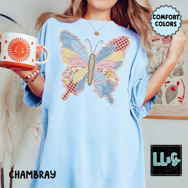 Peut inclure: T-shirt bleu clair avec un motif de papillon en patchwork. Le papillon pr&eacute;sente divers motifs de tissu roses, jaunes et rouges. Le mot "CHAMBRAY" est imprim&eacute; sur le t-shirt. Les mots "COMFORT COLORS" sont &eacute;galement visibles.