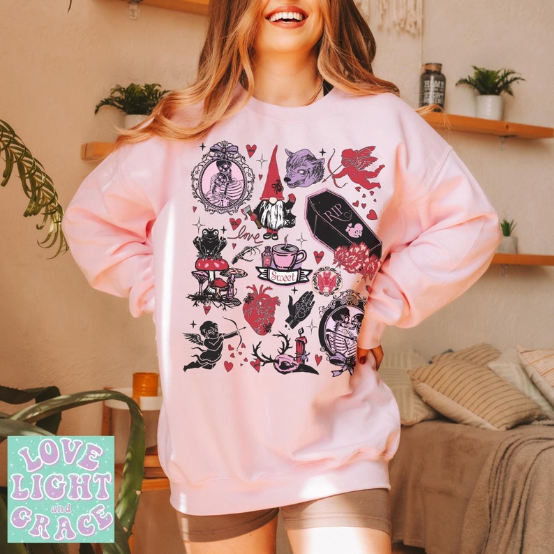 May include: A light pink sweatshirt featuring a collection of gothic-themed illustrations. The design includes a gnome, wolf, cupid, coffin, and other elements. The words "Love Light and Grace" are printed on the bottom left.