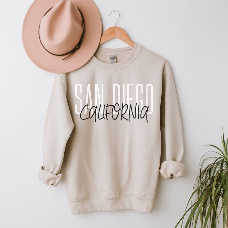 May include: A beige sweatshirt with the text "SAN DIEGO CALIFORNIA" printed in white and black on the front. The sweatshirt is hanging on a wooden hanger.