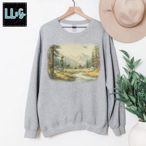 May include: A light gray sweatshirt featuring a landscape scene with an alien figure, trees, mountains, and a river. The image includes a flying saucer in the sky. The upper left corner has the text "LL&G" in a black box.