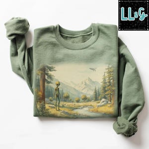 May include: Sage green sweatshirt featuring a landscape scene with an alien figure, trees, a river, and mountains. A flying saucer is in the sky. The sweatshirt has a crew neck and long sleeves. The brand logo "LL&G" is in the upper right corner.
