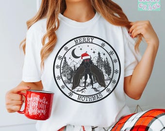 May include: White t-shirt with a circular graphic featuring a black moth wearing a Santa hat, the text "MERRY MOTHMAS", snowflakes, and a forest scene. A red mug with the text "Have yourself a Merry Little Christmas" is held in the hand.