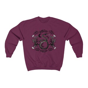 May include: A maroon sweatshirt with a black and white graphic print of a snake with a crescent moon and stars. The snake is surrounded by floral and leaf designs.