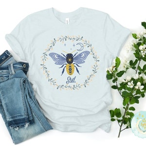May include: A light blue t-shirt with a floral wreath design featuring a stylized bee with a moon and stars. The text "Still" is printed below the bee.
