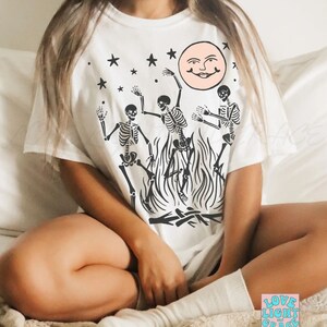 May include: White t-shirt with a graphic of three skeletons dancing around a bonfire with a smiling moon and stars in the background. The text "LOVE LIGHT and GRACE" is visible in the lower right corner of the image.