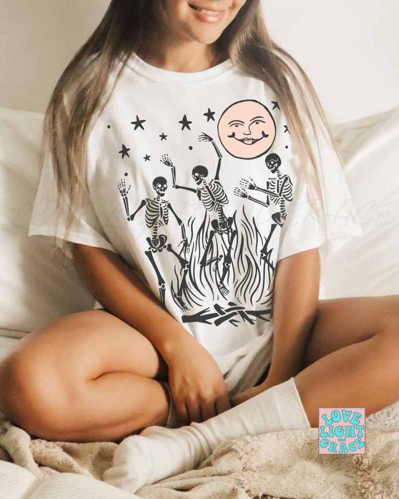 May include: White t-shirt with a graphic of three skeletons dancing around a bonfire with a smiling moon and stars in the background. The text "LOVE LIGHT and GRACE" is visible in the lower right corner of the image.