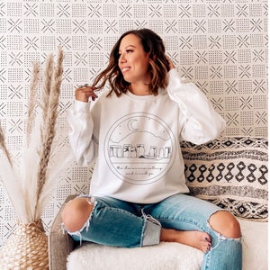 Puede incluir: Sudadera blanca con un dise&ntilde;o circular de Stonehenge, una luna creciente y estrellas. El texto "the stones are calling and I must go" est&aacute; debajo. La sudadera se combina con vaqueros azules rotos.