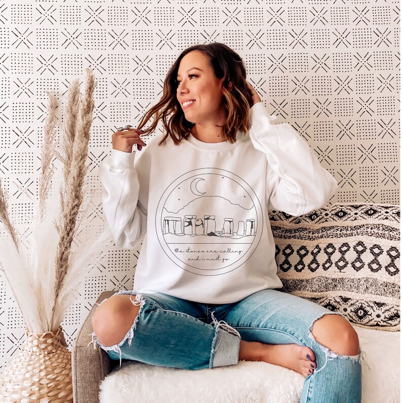 Puede incluir: Sudadera blanca con un dise&ntilde;o circular de Stonehenge, una luna creciente y estrellas. El texto "the stones are calling and I must go" est&aacute; debajo. La sudadera se combina con vaqueros azules rotos.