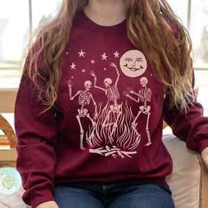 K&ouml;nnte beinhalten: Ein maroonfarbener Pullover mit einem Motiv von drei Skeletten, die um ein Lagerfeuer mit einem Vollmond im Hintergrund tanzen.