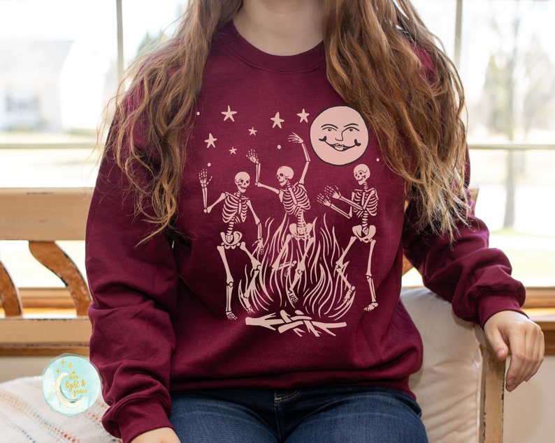 K&ouml;nnte beinhalten: Ein maroonfarbener Pullover mit einem Motiv von drei Skeletten, die um ein Lagerfeuer mit einem Vollmond im Hintergrund tanzen.