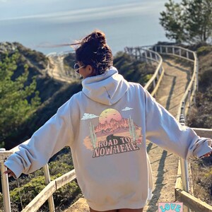 May include: A light grey hoodie featuring a graphic print of a desert landscape with the text "Road to Nowhere". The hoodie has a hood and a kangaroo pocket. The person wearing the hoodie is standing on a wooden walkway overlooking the sea.