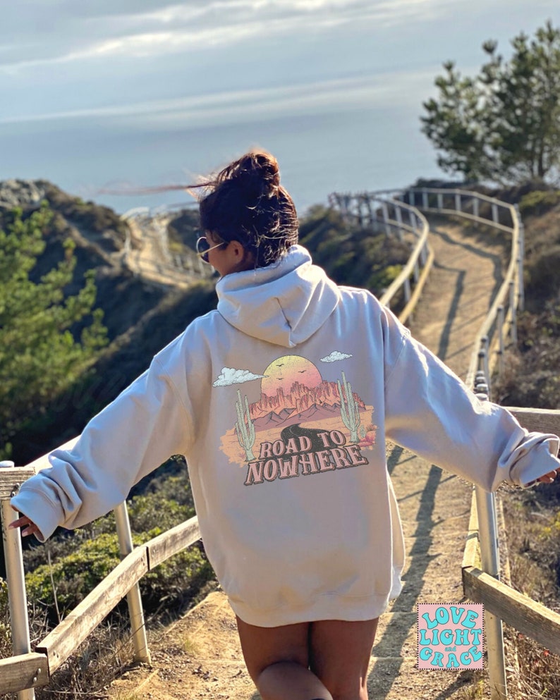 May include: A light grey hoodie featuring a graphic print of a desert landscape with the text "Road to Nowhere". The hoodie has a hood and a kangaroo pocket. The person wearing the hoodie is standing on a wooden walkway overlooking the sea.