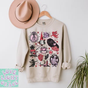 May include: A beige sweatshirt featuring a collection of whimsical illustrations in pink and black, including a gnome, a coffin, a heart, and a cherub. A pink hat hangs above the sweatshirt. The text "Love Light and Grace" is in the lower left corner.