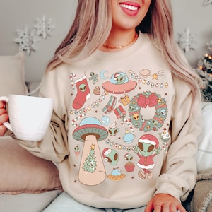 Könnte beinhalten: Ein beiger Pullover mit einem weihnachtlichen Design mit Außerirdischen, einem Weihnachtsbaum, einem Kranz und anderen festlichen Elementen. Das Design ist im Cartoon-Stil mit leuchtenden Farben gehalten.