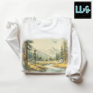 May include: White long-sleeve sweatshirt featuring a landscape scene with an alien figure, trees, mountains, and a flying saucer. The graphic is in muted tones of green, yellow, and blue. The upper right corner has the text "LL&G" in a black box.