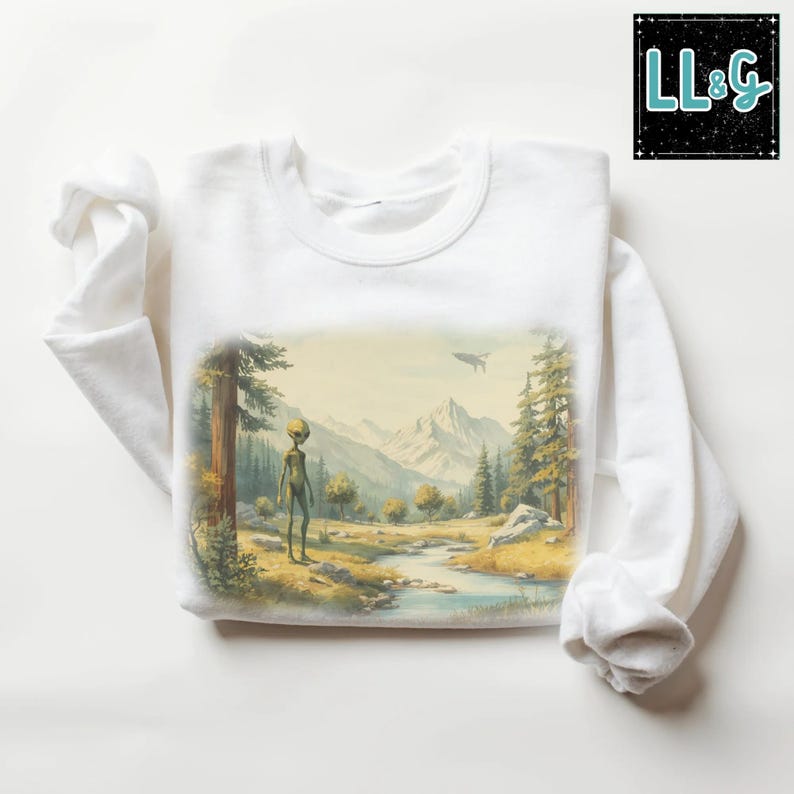 May include: White long-sleeve sweatshirt featuring a landscape scene with an alien figure, trees, mountains, and a flying saucer. The graphic is in muted tones of green, yellow, and blue. The upper right corner has the text "LL&G" in a black box.