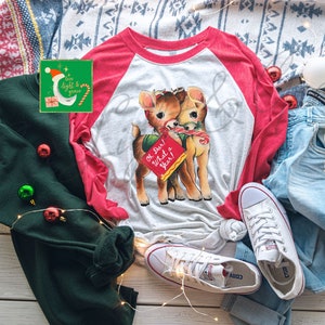 May include: A white and red long-sleeved shirt with a graphic of two deer holding a sign that says "Oh Dear! What a Year!" The shirt is on a white wooden surface with a green sweater, blue jeans, and white sneakers.