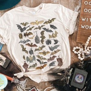 May include: A beige t-shirt featuring a print of bats in various poses. The text "Ooh Ooh, Witchy Woman" is visible on a sign in the background.