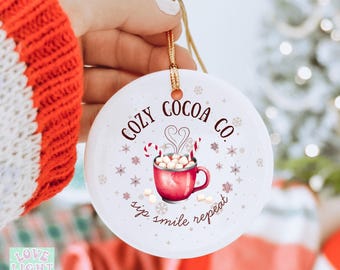 May include: A white ceramic Christmas ornament with the text "COZY COCOA CO." and a red mug of hot cocoa with marshmallows and candy canes. The ornament is held by a hand with a red sweater sleeve in the background.