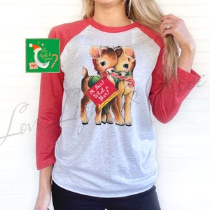 May include: A white and red baseball tee shirt with a vintage illustration of two deer holding a candy cane and a sign that says "Oh Deer! What a Year!"