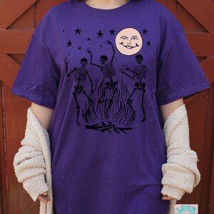 May include: Purple t-shirt with a black graphic of three skeletons dancing around a bonfire with a smiling moon and stars in the background.