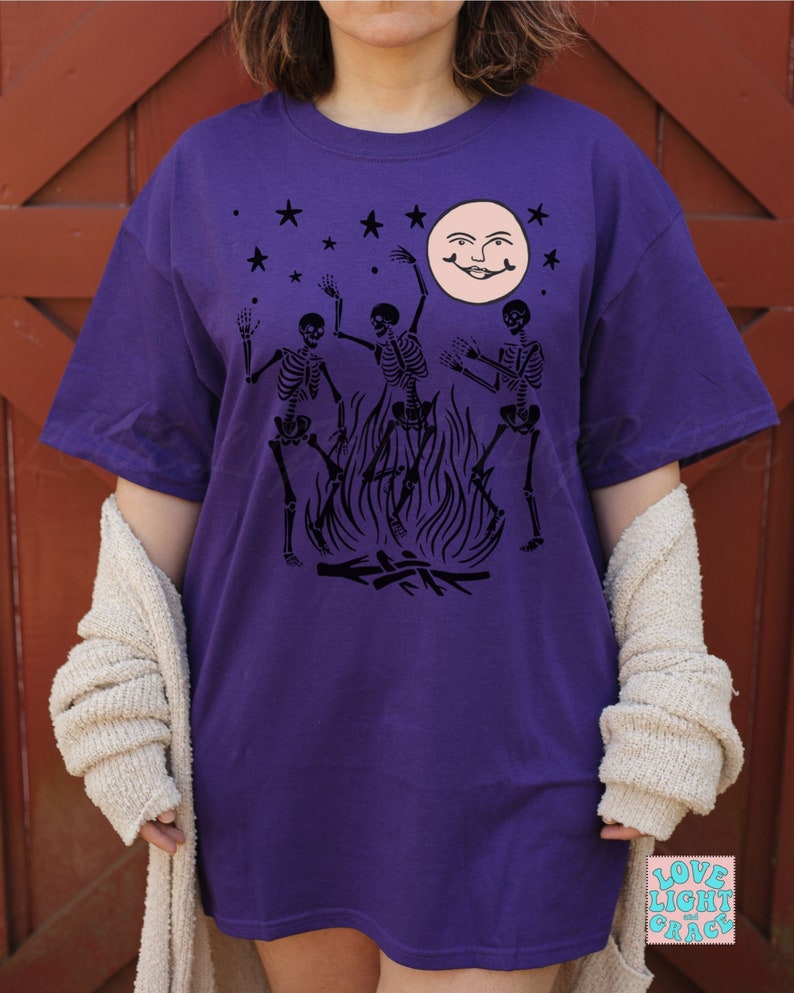 May include: Purple t-shirt with a black graphic of three skeletons dancing around a bonfire with a smiling moon and stars in the background.