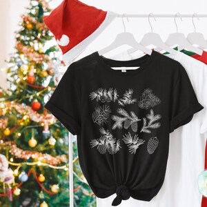 May include: Black t-shirt with a white design of pine branches and pine cones. The shirt is hanging on a hanger with other shirts.