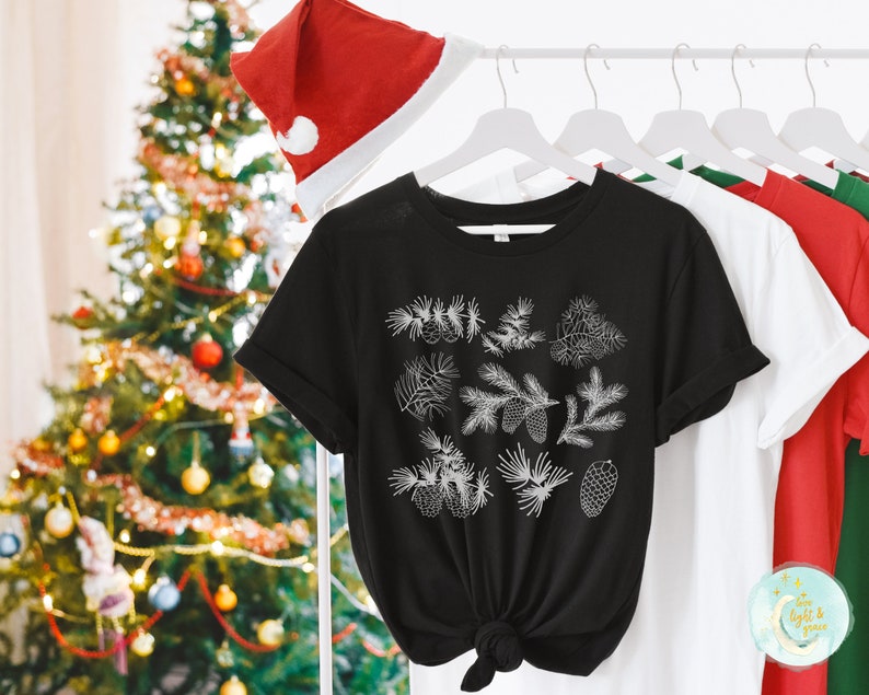 May include: Black t-shirt with a white design of pine branches and pine cones. The shirt is hanging on a hanger with other shirts.