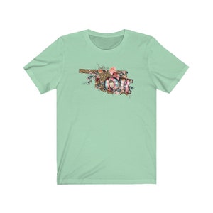 K&ouml;nnte beinhalten: Ein hellgr&uuml;nes T-Shirt mit einem floralen Design des Bundesstaates Oklahoma mit den Buchstaben "OK" in der Mitte. Die Buchstaben sind mit Lichtern und einem Leopardenmuster verziert.