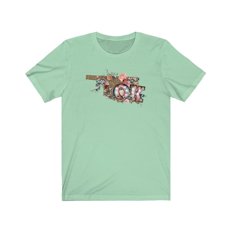 K&ouml;nnte beinhalten: Ein hellgr&uuml;nes T-Shirt mit einem floralen Design des Bundesstaates Oklahoma mit den Buchstaben "OK" in der Mitte. Die Buchstaben sind mit Lichtern und einem Leopardenmuster verziert.