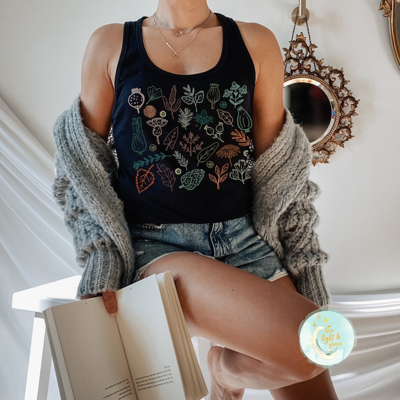 May include: A navy blue tank top featuring a botanical illustration of various leaves and flowers in shades of teal, orange, and green. The sleeveless top is worn with denim shorts and a gray cardigan.