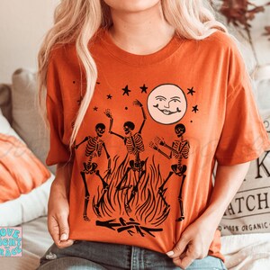 May include: A burnt orange t-shirt with a graphic of three skeletons dancing around a bonfire with a smiling moon in the background. The skeletons are black and the moon is white.