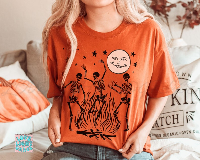May include: A burnt orange t-shirt with a graphic of three skeletons dancing around a bonfire with a smiling moon in the background. The skeletons are black and the moon is white.
