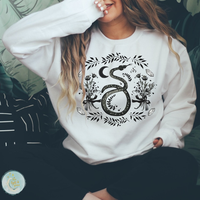 May include: White sweatshirt with a black and green snake design. The snake is curled around a crescent moon and surrounded by flowers, leaves, and crystals.
