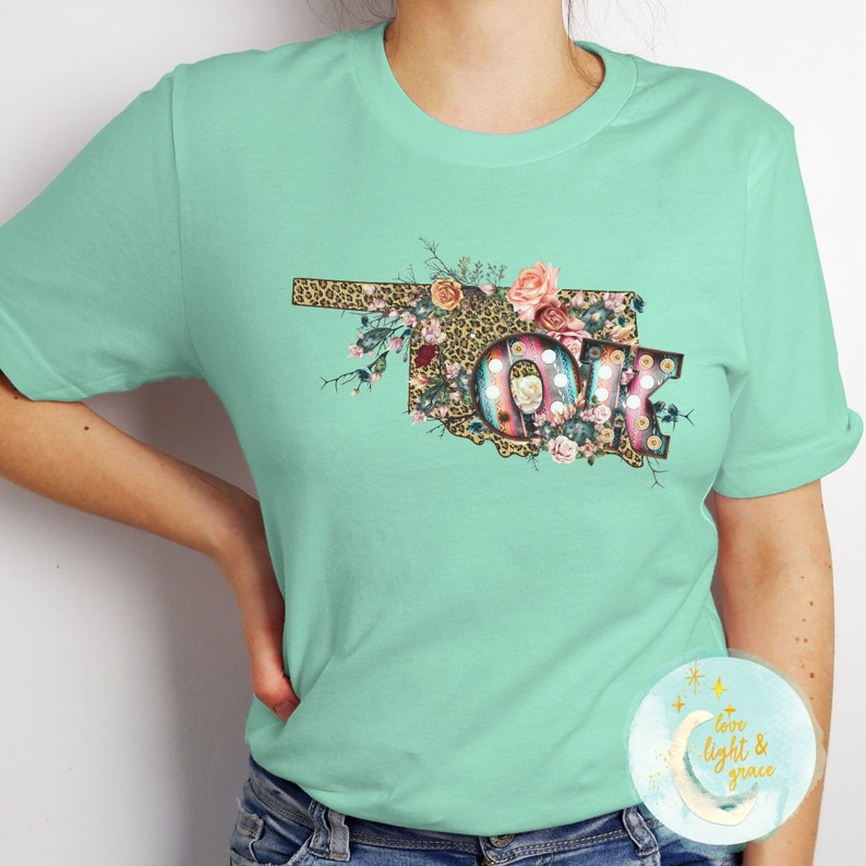 K&ouml;nnte beinhalten: Ein t&uuml;rkisfarbenes T-Shirt mit einem floralen Design, das den Bundesstaat Oklahoma und die Buchstaben "OK" im Vintage-Stil zeigt. Die Buchstaben sind in Gold umrandet und haben Gl&uuml;hbirnen. Das Design ist von Blumen und einem Leopardenmuster umgeben.