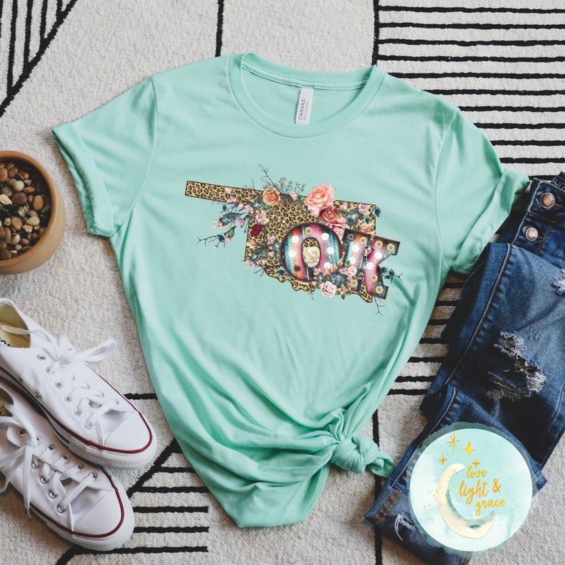 K&ouml;nnte beinhalten: Ein t&uuml;rkisfarbenes T-Shirt mit einem floralen Design, das den Bundesstaat Oklahoma und die Buchstaben "OK" im Stil einer Vintage-Leuchtreklame zeigt. Das Design beinhaltet Leopardenmuster, Blumen und Federn.