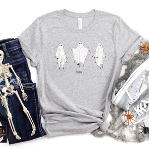 May include: A light gray t-shirt featuring three cartoon ghosts with the word "boo" printed below. The ghosts are white with black eyes and are standing. The shirt is paired with blue jeans and gray sneakers, with Halloween decorations.