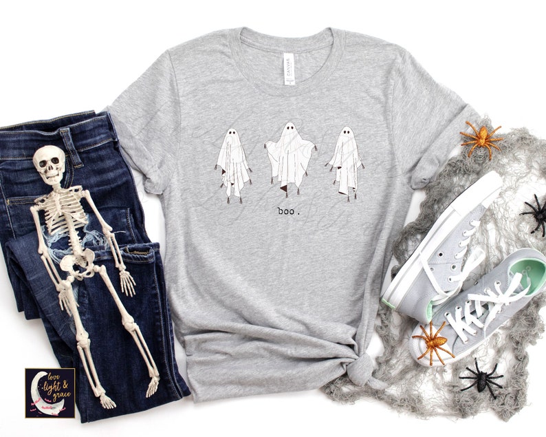May include: A light gray t-shirt featuring three cartoon ghosts with the word "boo" printed below. The ghosts are white with black eyes and are standing. The shirt is paired with blue jeans and gray sneakers, with Halloween decorations.