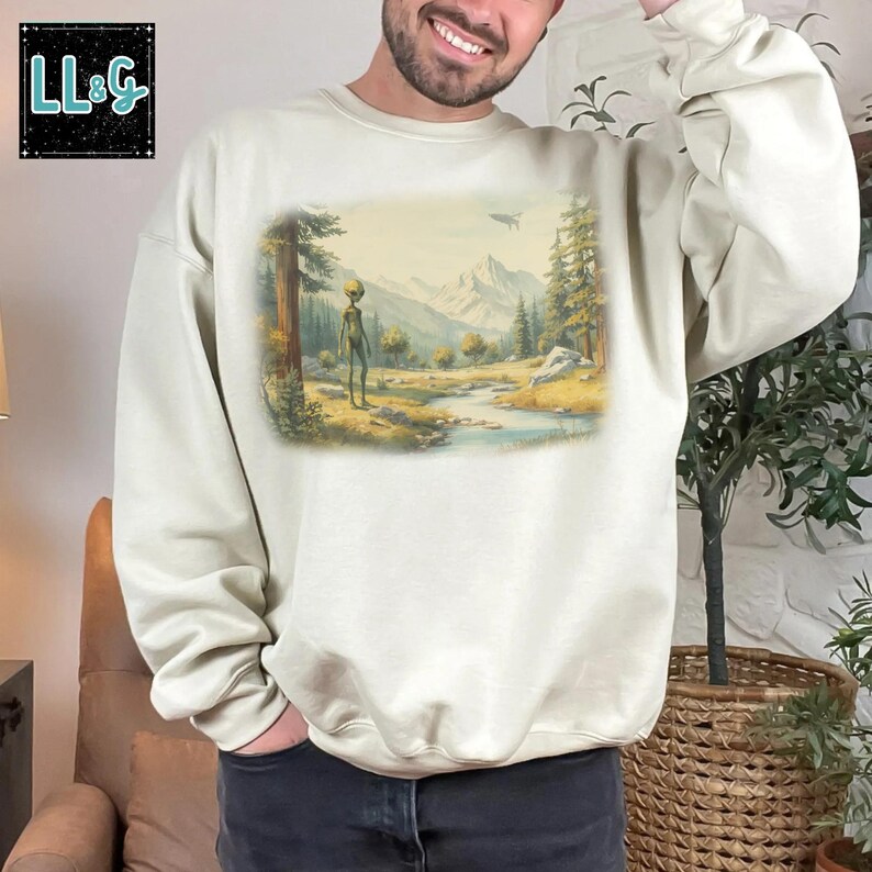 May include: Cream-colored sweatshirt featuring a landscape scene with an alien figure, mountains, and a river. The design includes a UFO in the sky. The upper left corner has the text "LL&G" in a black box. The sweatshirt is a casual, comfortable style.