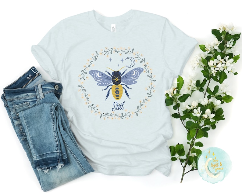 May include: A light blue t-shirt with a floral wreath design featuring a stylized bee with a moon and stars. The text "Still" is printed below the bee.