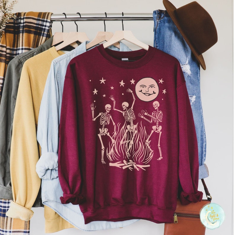 May include: A maroon sweatshirt with a graphic of three skeletons dancing around a bonfire with a smiling moon and stars in the background.