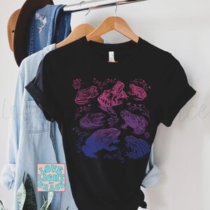 May include: A black t-shirt with a graphic print of frogs in shades of pink and blue. The frogs are arranged in a cluster and are surrounded by small floral designs. The text "LOVE LIGHT AND GRACE" is printed in pink and blue on a small square sticker in the bottom left corner of the image.