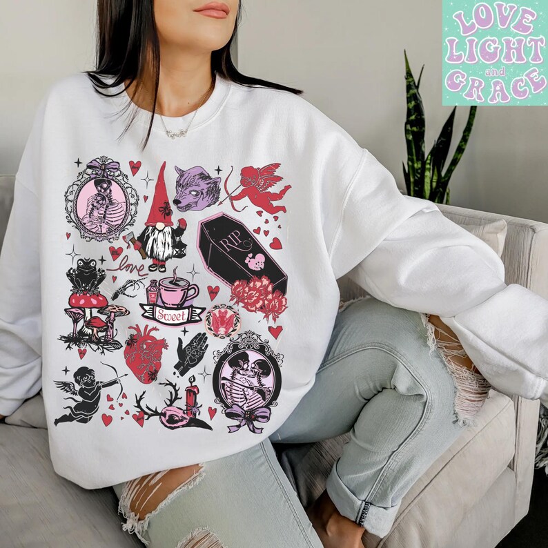 May include: White sweatshirt with a collection of whimsical illustrations in pink, red, and black. The design includes a skeleton couple, a gnome, a wolf, a cupid, a coffin, hearts, and the text "Sweet". The words "Love Light and Grace" are in the upper right corner.